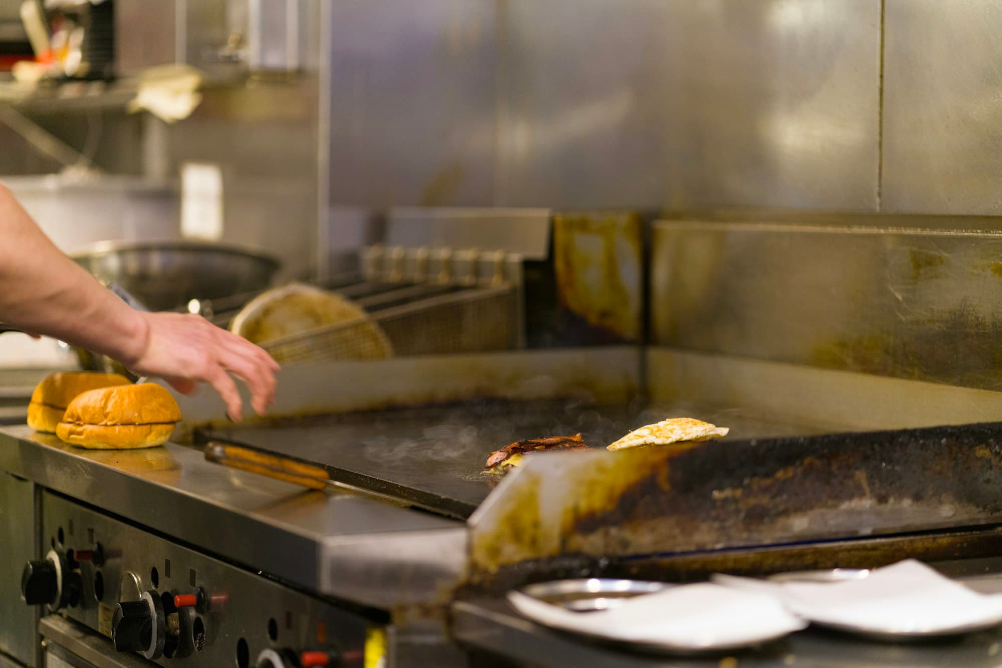 A cook working a flat-top grill with buns toasting, in a commercial kitchen.