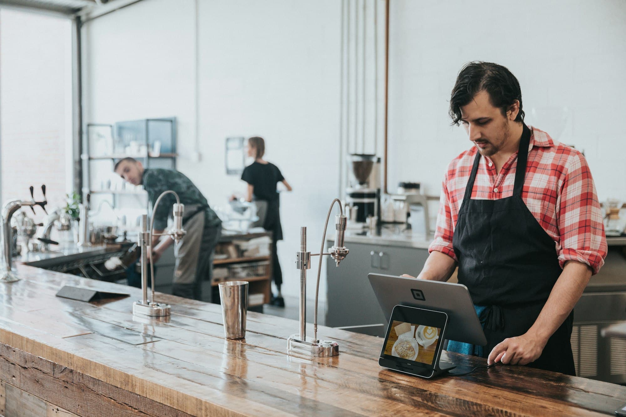 An owner in an apron checks a tablet at a café counter.